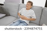 Small photo of Cute little blond boy suffering from stomach ache, sitting on home sofa with a worried face, touching unsettled stomach