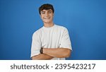 Small photo of Joyous, confident young hispanic teenager guy standing with crossed arms, spreading positive vibes, cheerfully smiling against isolated blue background.