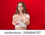 Small photo of Young caucasian woman standing over red background moving away hands palms showing refusal and denial with afraid and disgusting expression. stop and forbidden.