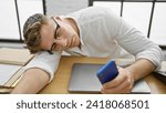 Small photo of Exhausted young caucasian businessman, smartphone in hand, loses battle with stressaE“ nobly crashes asleep on office table, job tedium takes its toll.