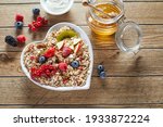 Top view of fresh honey and yogurt placed on wooden table near heart shaped bowl of muesli with pieces of fruits and berries in morning
