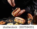 Small photo of Chef preparing a tasty hot dog with a frankfurter on a fresh roll adding extra trimmings sprinkling crispy bacon bits onto the pickles and sausage