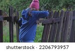 Small photo of Young Caucasian Girl Taking Shortcut Squeezing Through Hole in Damaged Wooden Fence and Trespassing Private Property