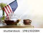 Glass teapot with tea brown cup on a wooden table. American flag in the background. Festive tea party.