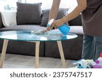 Small photo of Female housekeeper wearing apron and casual clothes standing at table with buckets of cleaning equipments holding alcohol spray and cloth to clean and wipe glass table in living room at home.
