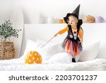 Small photo of toddler girl in witch costume with Magic wand and Halloween pumpkin on the bed the white interior. Adorable little blond girl ready for trick-or-treat. halloween, holiday and childhood concept.