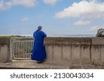 Small photo of A priest in a blue cassock looks into the distance at the sea. The clergyman prays to God on the sea coast. The concept of the frailty of human existence.