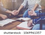 Christian small group holding hands and praying together around a wooden table with blurred open bible page in homeroom, devotional or prayer meeting concept