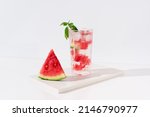 A glass of watermelon juice with pieces of watermelon and ice on light background