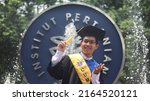 Small photo of BOGOR, INDONESIA - NOVEMBER 9, 2021: Young asian man taking a picture on university graduation day in IPB University campus landmarks "koin IPB". Bogor Agricultural University. Cumlaude predicate.