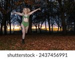 A young woman in a neon green leopard print swimsuit and fishnet tights poses with arms raised, against a twilight backdrop in a leaf-covered park.