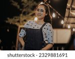 an Asian woman wearing glasses and wearing overalls is standing in front of the coffee shop terrace at night