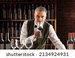 Small photo of Sommelier with a glass of wine. Examination of wine products. Restaurant staff, expert wine steward among shelves of wine bottles. Stylish middle-aged man with a grey beard.