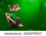 Irish girl holding big mugs with beer or ale sitting on cauldron with gold coins. Young red-haired woman as green Leprechaun elf. St. Patrick's Day party. Ireland National Independence Day March 17th.