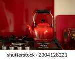 Small photo of Dark to light red ombre tea kettle with curved spout black knob, metal handle on stovetop front burner kitchen with deep red reflective backsplash boiling water cutting board scale fruit bowl lemon