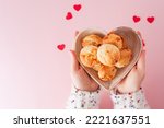A Girl`s hand holding one Brazilian cheese bread in top view in a soft pink background with red little hearts and a heart shaped bowl
