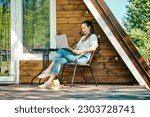 Young woman put laptop on her knees while resting on terrace of wooden a-frame cabin