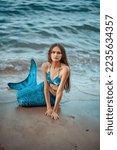 Small photo of Beautiful mermaid girl on the seashore. Mermaid on the ocean against the background of rocks. Mermaid with a blue tail on the beach. Mermaid on the background of the waves. Siren washed ashore.