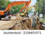 Small photo of Reconstruction of the underground sewerage system on a city street in spring. Two excavators are digging a deep ditch. Traffic has been halted on a section of the street. Road works.