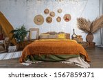Beautiful cozy bedroom with boho style interior, pillows, cushions, green plants in flower pot, bed and house decor on night table and wall
