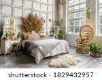 Side view of authentic cozy bedroom with interior design in boho chic style, decorative headboard over comfort bed, wicker armchair, houseplants near commode and mirror. Lovely room in classic house