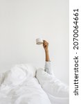 Small photo of Concept of early morning wake up. Vertical view dark skinned woman in pajamas on bed, wrapped in warmth cozy blanket, raise hand with coffee cup. Female girl holding mug against white copy space wall
