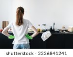 Small photo of back view of female in green rubber gloves standing and look at unclean tableware, glasses and dishware on worktop near sink, mess at home kitchen, housework concept