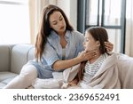 Small photo of Mother worries about her sick daughter, touching lymph nodes on neck while sitting on sofa near. Girl kid wrapped in plaid feeling weakness, fever and respiratory infection symptoms