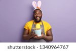 Small photo of Overemotive black man has broad toothy smile, appreciates Easter traditions, glad to meet family gather together, dressed in casual domestic clothes, holds yellow colored egg laid by rabbit.