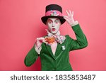 Small photo of Photo of shocked male has image of mysterious hatter from wonderland wears bright makeup poses with cup of tea dressed in aristocratic clothes isolated on pink background. Halloween party concept