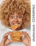 Small photo of Vertical image of surprised curly haired woman eats delicious sweet homemade belgian waffle enjoys favorite dessert has widen eyes poses against white background. High calories food. Diet breaking