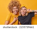 Small photo of Positive women have joyful expressions smile broadly make peace gesture laugh at something funny foolish around dressed casually isolated over vivid yellow background. Happy emotions concept