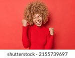 Small photo of Positive curly haired young woman makes yes gesture celebrates success feels overjoyed and triumphant wears casual turtleneck shakes fists exclaims from joy isolated over vivid red background