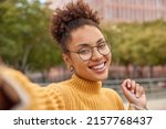 Small photo of Portrait of attractive smiling curly haired young woman in optical round glasses poses for selfie outdoors walks in urban setting during leisure time photographs herself has free day or weekend