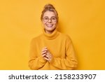 Small photo of Pretty blonde European woman smiles toothily keeps hands together feels satisfied wears round spectacles and casual loose jumper isolated over vivid yellow background. Happy emotions concept