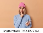 Small photo of Surprised mysterious young woman makes shush gesture tells secret holds mobile phone dressed in casual clothes asks to be quiet isolated over brown background. Hush dont say any word please.