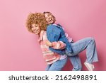 Small photo of Horizontal shot of cheerful best women friends give piggyback foolish around dressed in fashionable jacket and jeans have fun isolated over pink background. Friendship and free time concept.