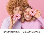Photo of two female best friends hold delicious doughnuts with marshmallow have sweet tooth enjoy eating tasty unhealthy food dressed casually isolated over pink background. Harmful nutrition