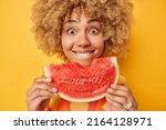 Close up shot of curly haired positive woman holds big slice of juicy watermelon enjoys eating her favorite summer fruit bites lips looks wondered isolated over yellow background. Mmm delicious