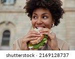 Pretty curly haired woman bites delicious sandwich poses outdoors at street looks away dressed casually has quick snack while walking outside being hungry. People lifestyle and fast food concept