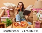 Small photo of Cheerful Asian woman enjoys eating pizza and watching film on portable tablet poses on floor around cardboard boxes rents new apartment. Relocation at house. Tenancy and real estate concept.