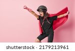 Small photo of Emotional woman superhero has extraordinary or superhuman powers ready to do heroic deeds makes flying gesture wears helmet eyemask rubber gloves cloak isolated on pink background fights against evil