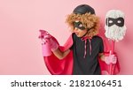 Small photo of Horizontal shot of scrupulous curly haired woman dressed in superhero costume sprays cleaning detergent holds mop ready to clean something dirty stands against pink background empty space aside