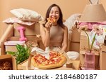 Happy young Asian woman eats appetizing pizza poses in messy room with cardboard boxes around relocates to new place of living isolated over beige background. People renting and mortgage concept