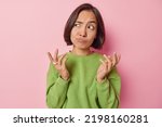 Small photo of Clueless puzzled Asian woman shrugs shoulders feels hesitant about something spreads hands sideways feels unaware dressed in casual green pullover isolated over pink background. So what to do