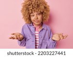Small photo of Clueless young woman with curly hair spreads palms feels unaware and questioned shrugs shoulders looks bewildered dressed in fashionable clothes isolated over pink background. Human perception