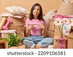 Thoughtful Asian woman sits crossed legs keeps index fimgers opposite each other thinks about relocation and changing place of living dressed in casual clothes surrpunded by cardboard boxes. Moving