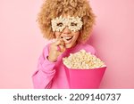 Indoor shot of surprised curly haired woman grabs delicious popcorn from paper bucket watches very intersting mobie excited by breathtaking scene dressed casually isolated over pink background
