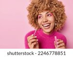 Small photo of Positive dreamy woman with curly hair applies lip gloss wears vivid makeup smiles broadly has perfect white teeth wears casual pink jumper poses indoor undergoes beauty procedures wants to look nice