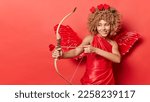 Small photo of Horizontal shot of cheerful female cupid shoots arrow targets on something wears red dress with wings behind back looks mysteriously somewhere isolated over red background copy space for your text